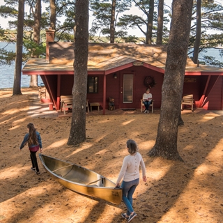 Two people carry a canoe while another person reads a book in front of a cabin in Wisconsin Dells.