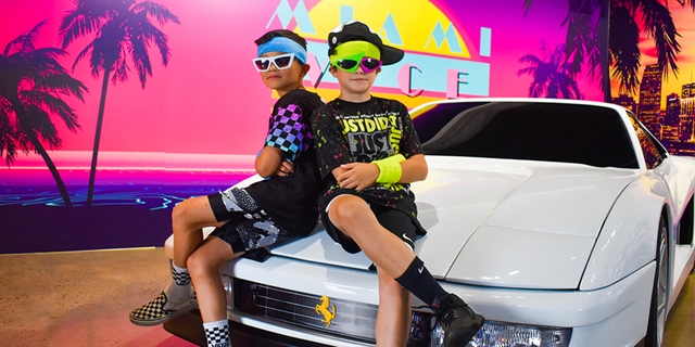 Two kids in 1980's attire pose on a vintage Ferrari.
