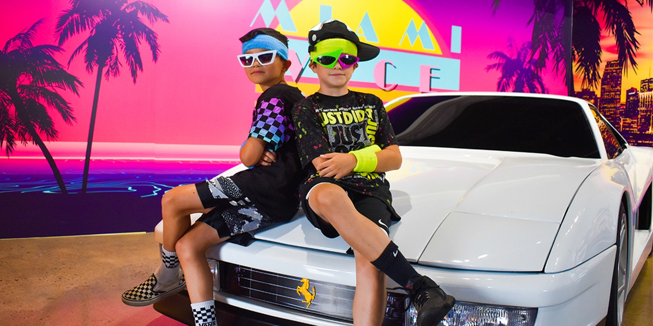 Two kids in 1980's attire pose on a vintage Ferrari.